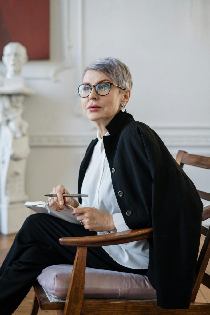 Senior woman with glasses sitting in a stylish office setting, holding a notebook and pen, signifying professionalism.